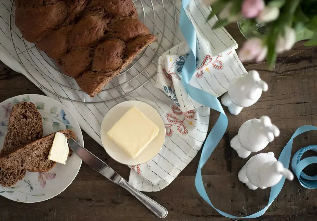 Frischer Hefezopf auf einem Drahtgestell, umgeben von Butter, Blumen und blauen Bändern auf einem Holztisch.