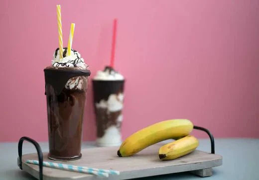 Veganer Schokoladen-Bananen-Milchshake mit Sahne und zwei Trinkhalmen vor pinkem Hintergrund, daneben zwei Bananen.