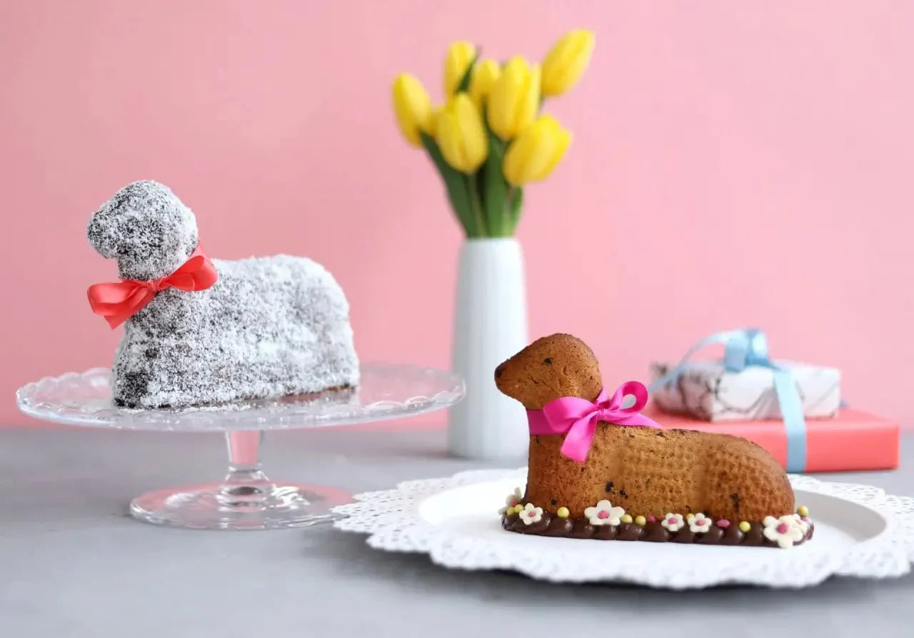 Zwei Osterlamm-Kuchen auf Tellern vor rosa Hintergrund, dekoriert mit Schleifen und Blumen, gelbe Tulpen im Hintergrund.