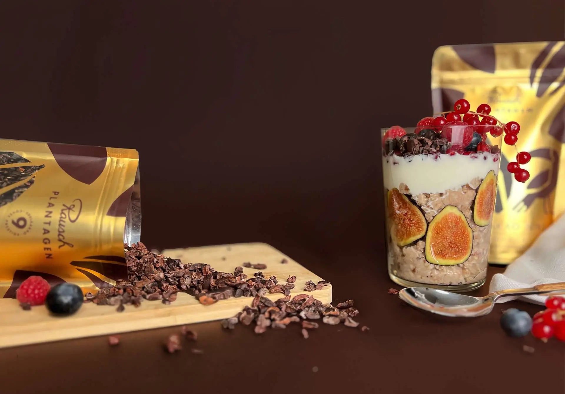 Kakao-Nibs auf einem Holzbrett und ein Dessertglas mit Haferbrei, Früchten und Nibs, vor einer braunen Hintergrundkulisse.