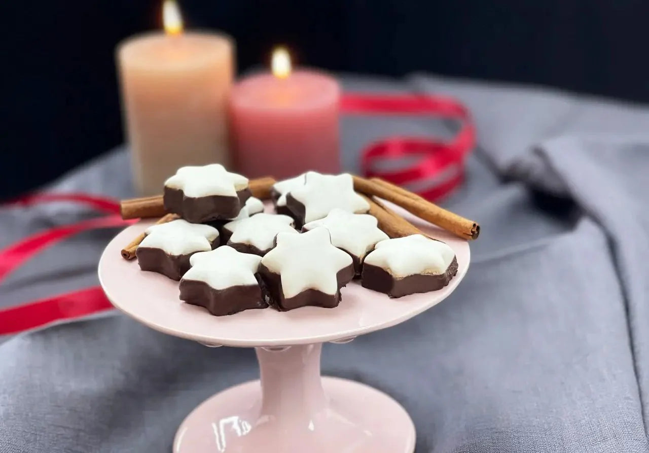 Weiße Zimtsterne auf Tortenplatte mit Kerzen und Zimtstangen, weihnachtliche Dekoration im Hintergrund.