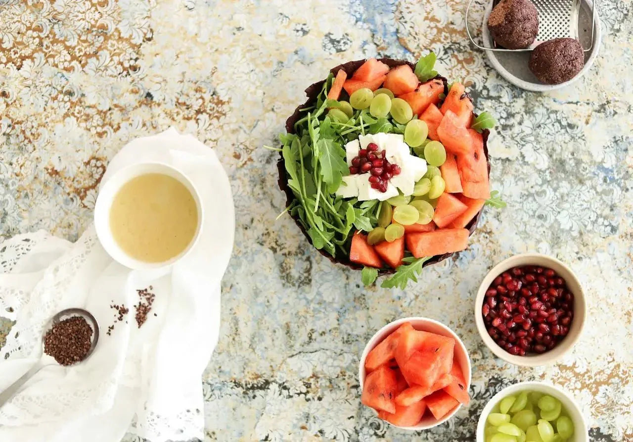 Bunter Sommersalat mit Melone, Weintrauben, Rucola und Kakaonibs, daneben Schalen mit Zutaten und Dressing.