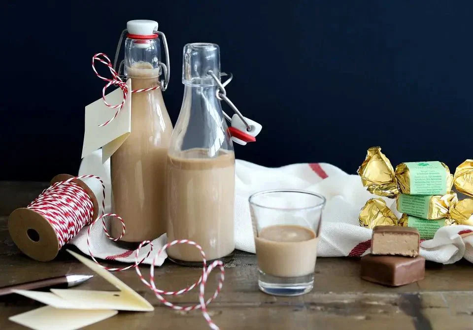 Selbstgemachter Nougatlikör in Flaschen mit Glas, Geschenkband und Schokolade auf einem Holztisch.