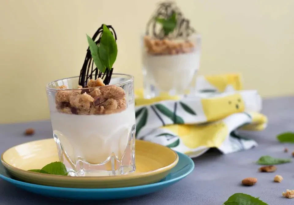 Panna Cotta im Glas mit Keksstreuseln und Schokodekor, verziert mit Zitronenverbene, auf buntem Teller präsentiert.