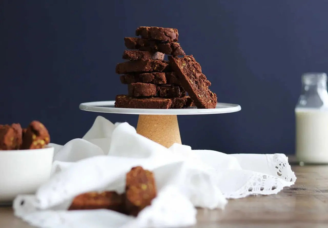 Ein Stapel Pistazien-Cantuccini mit Edelkakao-Schokolade auf einer weißen Platte, im Hintergrund ein Glas Milch.