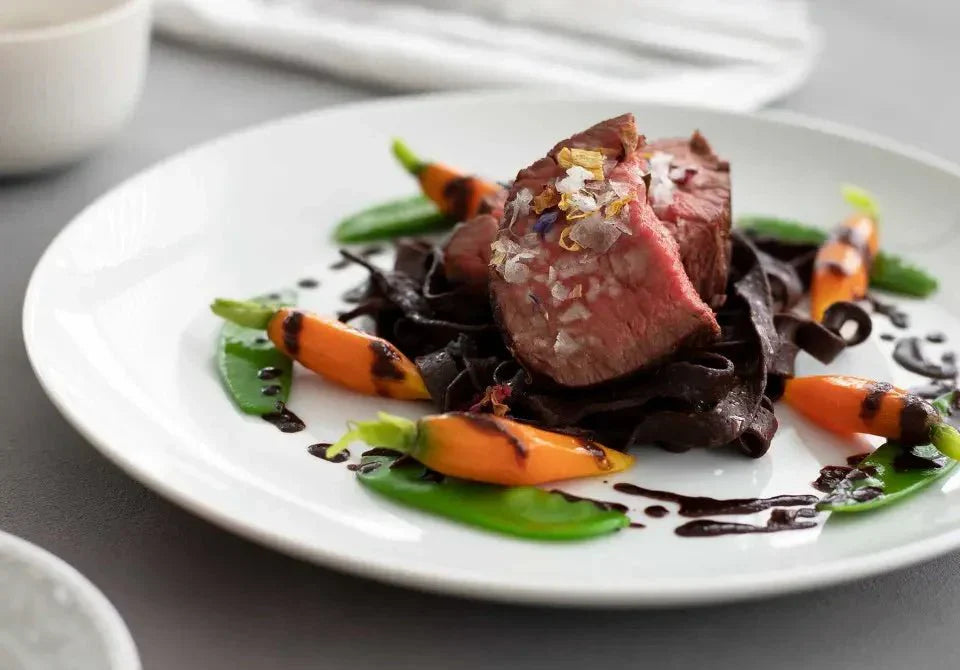 Zartes Rinderfilet auf Schokoladen-Nudeln mit Schokoladen-Portwein-Jus, garniert mit Babygemüse auf weißem Teller.