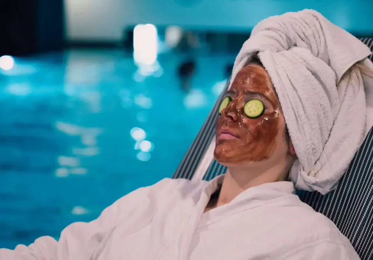 Person mit Schokoladenmaske und Gurkenscheiben auf den Augen entspannt am Pool, in einem Bademantel.