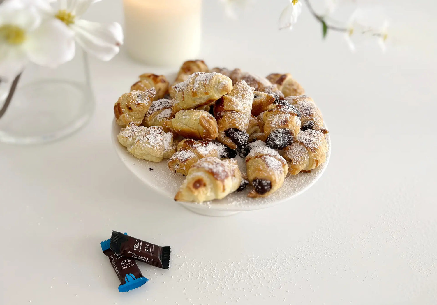 Frische, goldbraune Schokocroissants mit Puderzucker auf einer Tortenplatte, dekoriert mit Blüten.