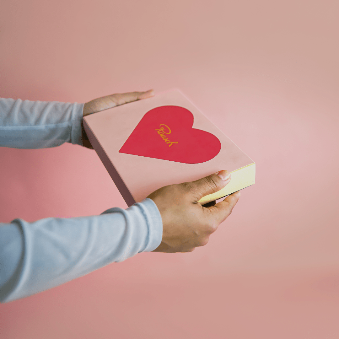 Zwei Hände halten die Pralineneschachtel mit Herzmotiv vor rosafarbenen Hintergrund.